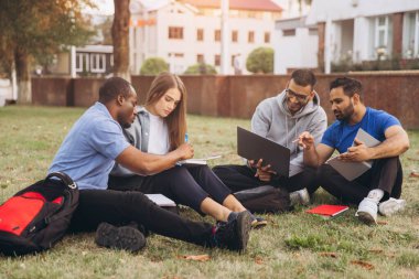 Kampüste birlikte çalışan bir grup üniversite öğrencisi. Çimlerin üzerinde oturuyorlar, gülümsüyorlar, fikirleri paylaşıyorlar ve dizüstü bilgisayar ve defterler kullanıyorlar..