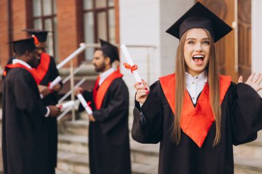 Mutlu genç bir kadın arkadaşlarıyla birlikte mezun oluyor, akademik elbiseler ve havan mermileriyle dışarıda kutlama yapıyor, özel bir günde neşe ve başarıyı ifade ediyor..