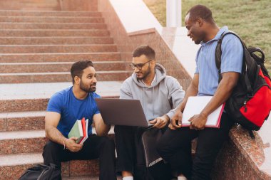 Üç farklı öğrenci kampüs basamaklarında oturmuş dışarıda ders çalışırken ve takım çalışması yaparken mutlu bir şekilde iş birliği yapıyorlar..