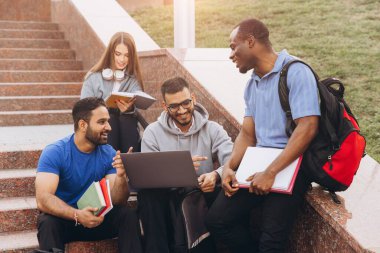 Bir proje için dizüstü bilgisayar ve kitaplar kullanarak kampüs basamaklarında tartışma ve işbirliği yapan çeşitli öğrenci grupları.