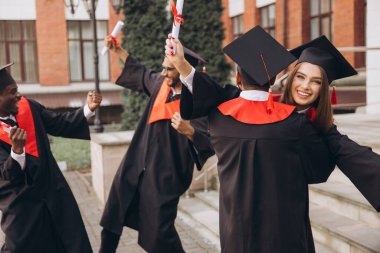 Heyecanlı mezunlar, başarılarından mutluluk ve gururla diplomalarını taşırken, başlık ve cübbe giyerek birlikte dışarıda kutlama yapıyorlar..