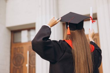 Arka Bakış: Başlık ve cübbeli genç bir bayan üniversite binasının önünde gururla diploma tutuyor, akademik başarıyı ve yeni başlangıçları kutluyor..