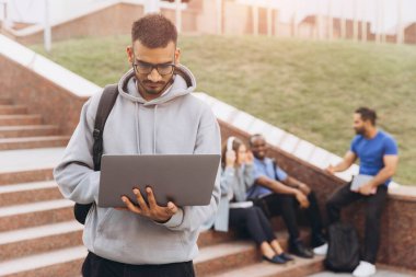 Kapüşonlu genç Hintli bir adam, güneşli bir günde arka planda bir grup arkadaş sosyalleşirken dışarıda dizüstü bilgisayarına konsantre oluyor..