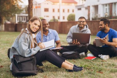 Mutlu ve çeşitli üniversite öğrencileri kampüste birlikte okuyorlar. Dışarıda dizüstü bilgisayarlar ve dizüstü bilgisayarlarla toplanıyorlar, rahat bir öğrenme ortamında takım çalışmalarını ve arkadaşlığı teşvik ediyorlar..