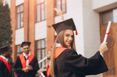 Mutlu genç bir kadın arkadaşlarıyla birlikte mezun oluyor, akademik elbiseler ve havan mermileriyle dışarıda kutlama yapıyor, özel bir günde neşe ve başarıyı ifade ediyor..