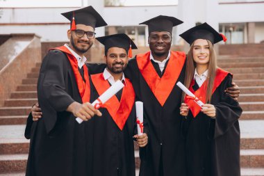 Şapka ve cübbe giymiş, gururla diploma taşıyan neşeli bir grup mezun. Mezuniyet gününde akademik başarılarını ve başarılarını kutluyorlar..