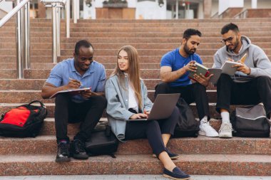 Üniversite basamaklarında öğrenim gören, dizüstü bilgisayar ve not defteri kullanarak bilgi paylaşan çeşitli bir öğrenci grubu. Açık hava akademik işbirliği.