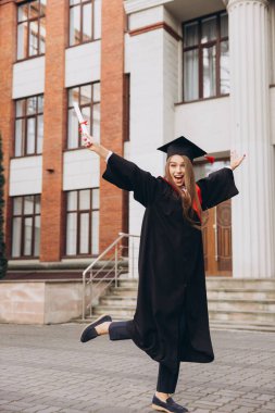 Mezuniyet elbisesi içindeki mutlu genç bir kadın, tarihi bir üniversite binasının önünde başarı ve başarı anısına diplomasını tutarak heyecanla atlıyor..