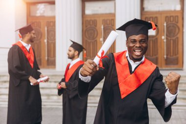 Bir grup mutlu mezun, üniversite binasının önünde ellerinde diplomalarla başarılarını kutluyorlar. Mutluluk ve başarının ifadeleri.