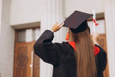 Arka Bakış: Başlık ve cübbeli genç bir bayan üniversite binasının önünde gururla diploma tutuyor, akademik başarıyı ve yeni başlangıçları kutluyor..