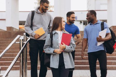 Sırt çantaları ve kitaplar taşıyan, arkadaşlıklarını ve akademik yaşamlarını sergileyen, gülümseyen ve kampüs merdivenlerinden inen dört üniversite öğrencisinden oluşan çeşitli bir grup..