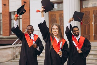 Üç farklı mezun dışarıda başarılarını kutluyor, ellerinde şapkalar ve diplomalar var. Onların ifadeleri mezuniyet gününde neşe ve başarıyı yansıtır..