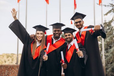 Çeşitli mezunlardan oluşan mutlu bir grup akademik başarılarını kutluyor, diplomaları heyecan ve gururla açık havada tutuyorlar..