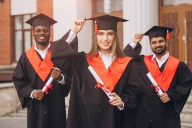 Çeşitli mezunlardan oluşan neşeli bir grup, başarılarını dışarıda kutluyor, ellerinde diplomalar ve kırmızı aksanlı geleneksel kepler ve elbiseler var..