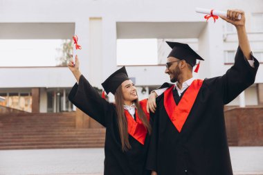 İki mutlu mezun genç adam ve kadın şapkalı ve cüppeli diplomalarla kutlama yapıyorlar. Üniversite binasının dışında duruyorlar, akademik yolculuklarında gurur ve başarı belirtiyorlar..