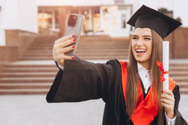 Şapka ve cübbe giymiş genç bir bayan elinde diplomayla kutlama selfie 'si çekiyor, dışarıda neşe ve başarı ifade ediyor..