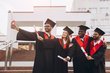 Akademik şapkalar ve elbiseler giyen, diplomalarını tutan, neşe ve başarılarını ifade eden çeşitli gülümseyen mezunlardan oluşan bir grup başarılarını dışarıda kutluyor..