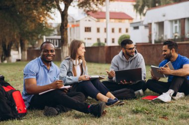 Üniversite öğrencilerinden oluşan çeşitli bir grup kampüsün bahçesinde ders çalışmaktan zevk alıyor, genç yetişkinler arasında işbirliği, takım çalışması ve arkadaşlık sergiliyorlar..