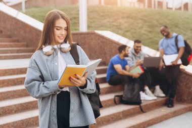 Kulaklığı ve not defteri olan neşeli bir öğrenci kampüs merdivenlerinde arkadaşlarıyla birlikte duruyor. Akademik enerji ve gençlik yoldaşlığı.