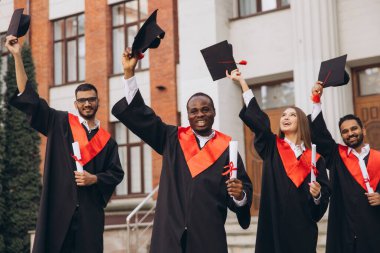 Çeşitli mezunlar başarılarını sevinçle kutluyor, diploma ve şapkalarını açık havada tutuyorlar. Başarı, çeşitlilik ve eğitimi temsil eder.