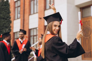 Mutlu genç bir kadın arkadaşlarıyla birlikte mezun oluyor, akademik elbiseler ve havan mermileriyle dışarıda kutlama yapıyor, özel bir günde neşe ve başarıyı ifade ediyor..