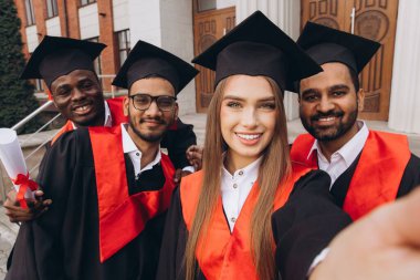 Bir grup farklı üniversite mezunu üniversite binasının önünde mezuniyet kepleri ve cübbeleriyle selfie çekerken neşeyle gülümsüyor..