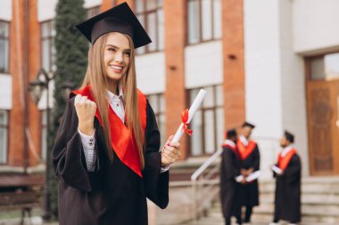 Mezuniyet şapkası ve cübbesiyle gülümseyen genç bir kadın, elinde diplomasıyla, başarıyı ve başarıyı sembolize ediyor, arka planda güneşli bir kampüs gününde sınıf arkadaşları var..