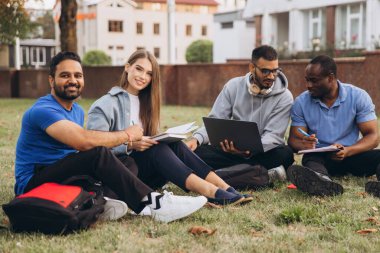 Genç üniversite öğrencileri kampüsün bahçesinde oturup bir çalışma projesinde işbirliği yapıyorlar. Bağlı ve odaklanmış halde fikirleri paylaşıyor ve doğal bir ortamda birlikte çalışıyorlar..