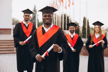 Geleneksel kep ve cübbe giymiş bir grup mutlu mezun akademik başarılarını ellerinde diplomalarla, gurur ve başarı dolu bir şekilde dışarıda kutluyorlar..