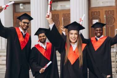 Çeşitli genç mezunlar ellerinde diplomalarla kutlama yapıyorlar. Geleneksel kepler ve cübbeler giyip bir üniversite binasının önünde duruyorlar..