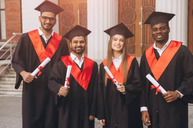 Başlık ve cübbe giymiş dört farklı mezun ellerinde diplomalarla bir üniversite binasının dışında akademik başarılarını kutluyorlar..