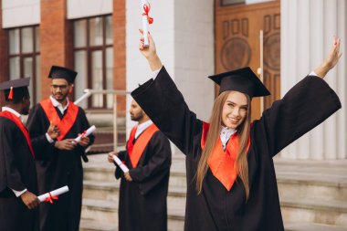 Neşeli mezunlar üniversite binasının dışında, kep ve cübbe giyerek, diploma taşıyarak, eğitimde başarı ve başarı göstererek kutlama yapıyorlar..