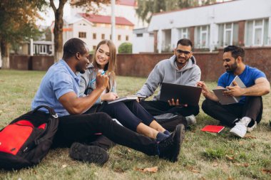 Kampüste birlikte çalışan bir grup üniversite öğrencisi. Çimlerin üzerinde oturuyorlar, gülümsüyorlar, fikirleri paylaşıyorlar ve dizüstü bilgisayar ve defterler kullanıyorlar..