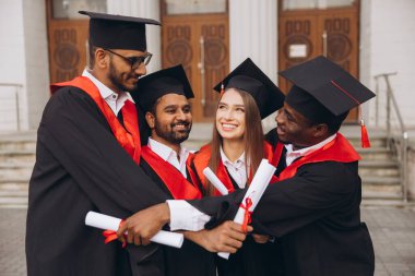 Çeşitli mezunlardan oluşan neşeli bir grup, akademik önlük ve şapkalardaki başarılarını kutluyor, diplomalarını bir üniversite binasının önünde açık havada tutuyorlar..