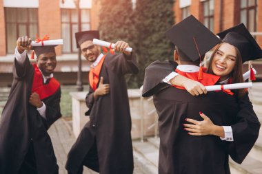 Çeşitli mezunlardan oluşan neşeli bir grup kep ve cübbe başarılarını kutluyor, diploma tutuyor ve birbirlerine sarılıyorlar. Bir üniversite binasının önünde yakalanmış..