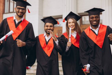 Dört mutlu mezundan oluşan çeşitli bir grup, bir üniversite binasının önünde diplomalarını tutarak kep ve cübbe giymedeki başarılarını kutluyor..