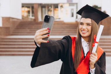 Şapka ve cübbe giymiş genç bir bayan elinde diplomayla kutlama selfie 'si çekiyor, dışarıda neşe ve başarı ifade ediyor..