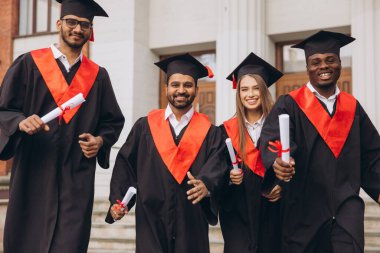 Dört mutlu mezundan oluşan çeşitli bir grup, bir üniversite binasının önünde diplomalarını tutarak kep ve cübbe giymedeki başarılarını kutluyor..