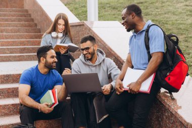 Bir proje için dizüstü bilgisayar ve kitaplar kullanarak kampüs basamaklarında tartışma ve işbirliği yapan çeşitli öğrenci grupları.