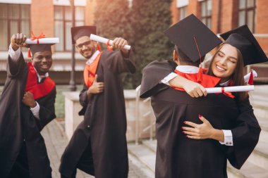 Çeşitli mezunlardan oluşan neşeli bir grup kep ve cübbe başarılarını kutluyor, diploma tutuyor ve birbirlerine sarılıyorlar. Bir üniversite binasının önünde yakalanmış..