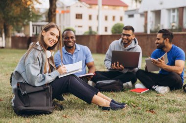 Mutlu ve çeşitli üniversite öğrencileri kampüste birlikte okuyorlar. Dışarıda dizüstü bilgisayarlar ve dizüstü bilgisayarlarla toplanıyorlar, rahat bir öğrenme ortamında takım çalışmalarını ve arkadaşlığı teşvik ediyorlar..