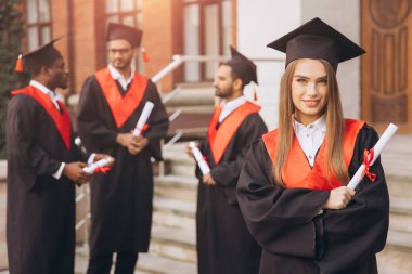 Mutlu genç bir kadın arkadaşlarıyla birlikte mezun oluyor, akademik elbiseler ve havan mermileriyle dışarıda kutlama yapıyor, özel bir günde neşe ve başarıyı ifade ediyor..