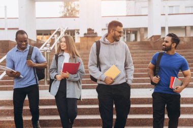 Üniversite öğrencilerinden oluşan çeşitli bir grup kampüs merdivenlerinde gülümseyip defterlerini taşıyarak güneşli bir günün tadını çıkarıyorlar. Dostluğu, eğitimi ve çok kültürlü çeşitliliği temsil eder.