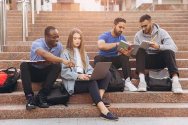 Üniversite kampüs basamaklarında dizüstü bilgisayar ve kitap kullanarak birlikte çalışan çeşitli bir öğrenci grubu. Açık havada bağlı ve işbirlikçi öğrenme atmosferi.