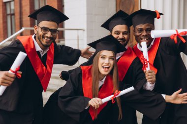 Çeşitli mezunlardan oluşan neşeli bir grup başarılarını kutluyor ve ellerinde diplomalar var. Mezuniyet gününde açık havada başarı ve başarının neşeli ifadesi..