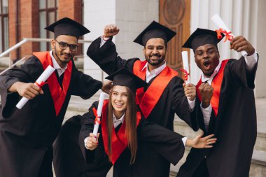Dört farklı üniversite mezunundan oluşan neşeli bir grup. Başlık ve cübbe giyip, diplomalarını tutarken başarılarını kutluyorlar ve mutlu bir şekilde poz veriyorlar..