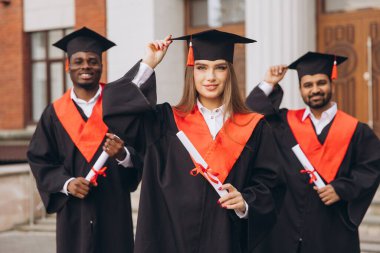 Çeşitli mezunlardan oluşan neşeli bir grup, başarılarını dışarıda kutluyor, ellerinde diplomalar ve kırmızı aksanlı geleneksel kepler ve elbiseler var..