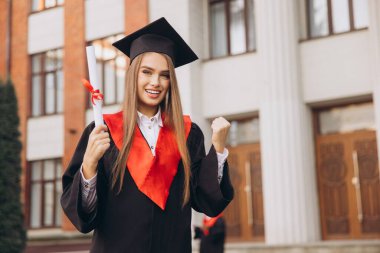 Kepli ve cüppeli mutlu bir mezun, büyük bir üniversite binasının önünde diplomasıyla sevinçle kutluyor. Başarının ve başarının özünü yakalamak.