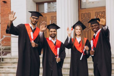 Farklı mezunlardan oluşan bir grubun dışarıda başarılarını kutladıkları, kep ve cübbe giydikleri ve diploma taşıdıkları mutlu bir an..
