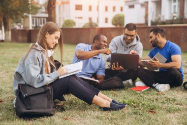 Mutlu ve çeşitli üniversite öğrencileri kampüste birlikte okuyorlar. Dışarıda dizüstü bilgisayarlar ve dizüstü bilgisayarlarla toplanıyorlar, rahat bir öğrenme ortamında takım çalışmalarını ve arkadaşlığı teşvik ediyorlar..